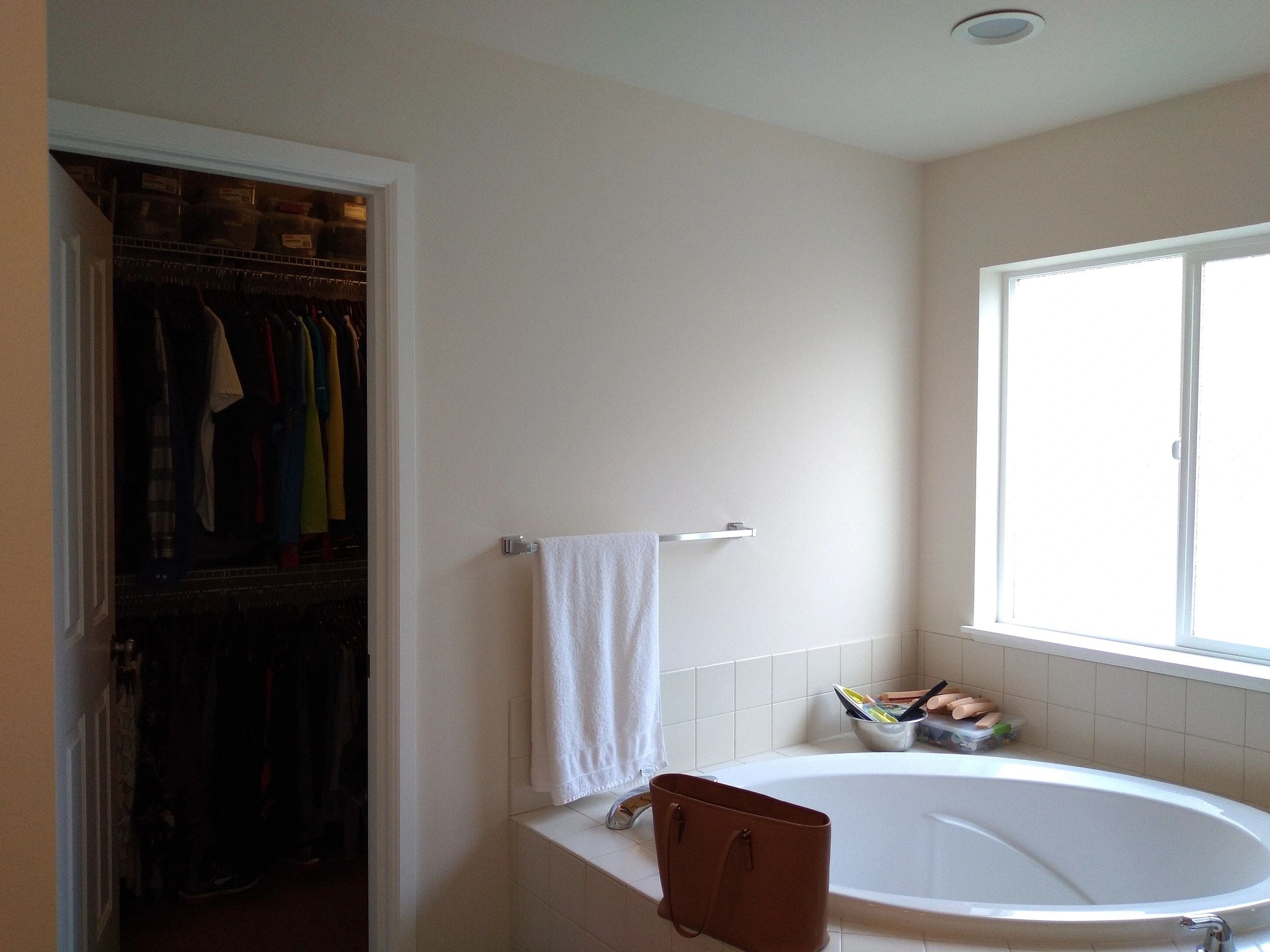 A bathroom with a large bathtub and a window next to an open closet filled with clothes and accessories.